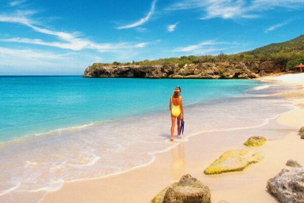 A woman in a yellow swimsuit walking on a beach with clear blue water and a rocky outcrop in the background.