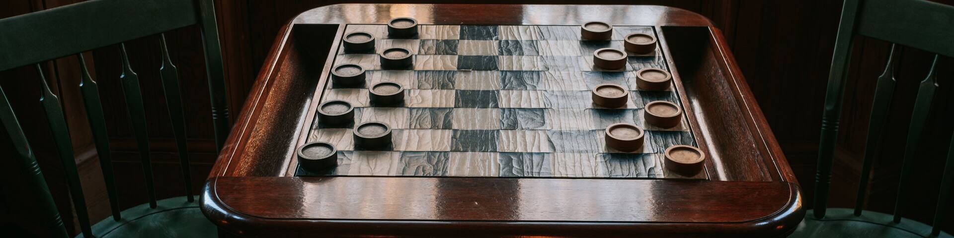 A wooden chess table with a checkered board and pieces, surrounded by wooden chairs.