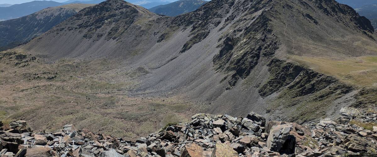 A rugged mountain landscape with rocky outcrops and distant snow-capped peaks under a clear sky.