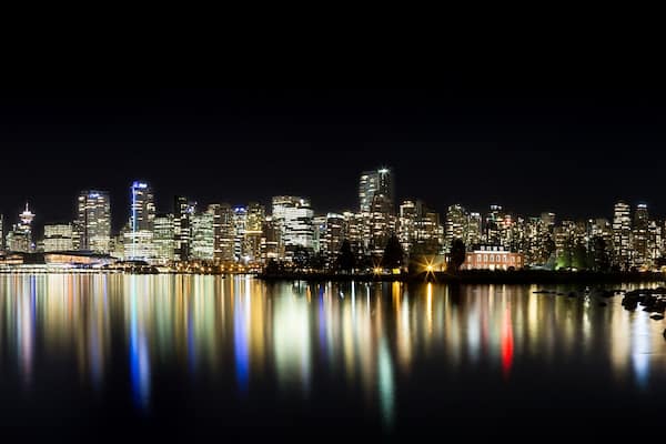 La silhouette d’une ville illuminée la nuit se reflète sur des eaux calmes, avec un pont imposant et une rangée de bâtiments aux lumières vives.