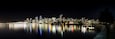 A nighttime city skyline reflected in calm waters, with a prominent bridge and a row of brightly lit buildings.