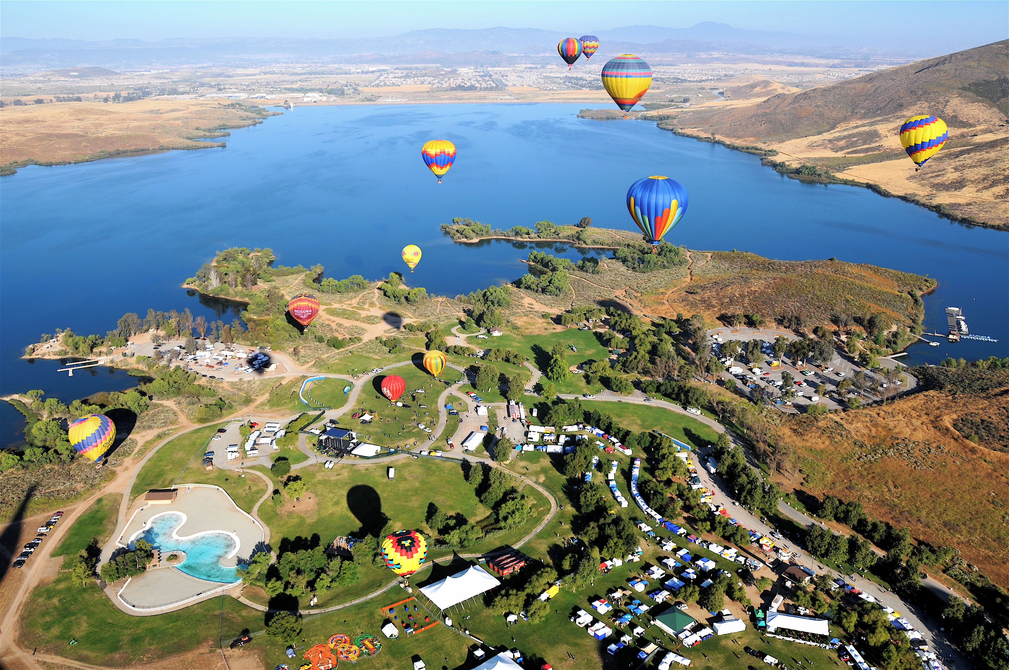 An aerial view of a hot air balloon festival over a lake with multiple balloons, a parking area, and surrounding greenery.