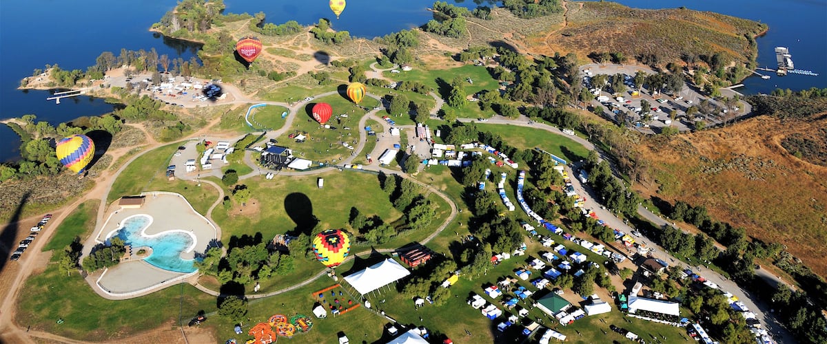 An aerial view of a hot air balloon festival over a lake with multiple balloons, a parking area, and surrounding greenery.