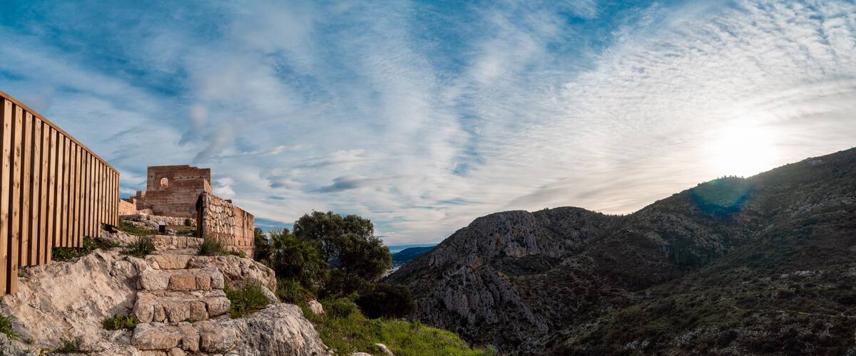 A historic stone structure with a wooden fence, set against a mountainous backdrop and a partly cloudy sky.