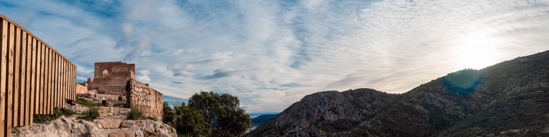 A historic stone structure with a wooden fence, set against a mountainous backdrop and a partly cloudy sky.