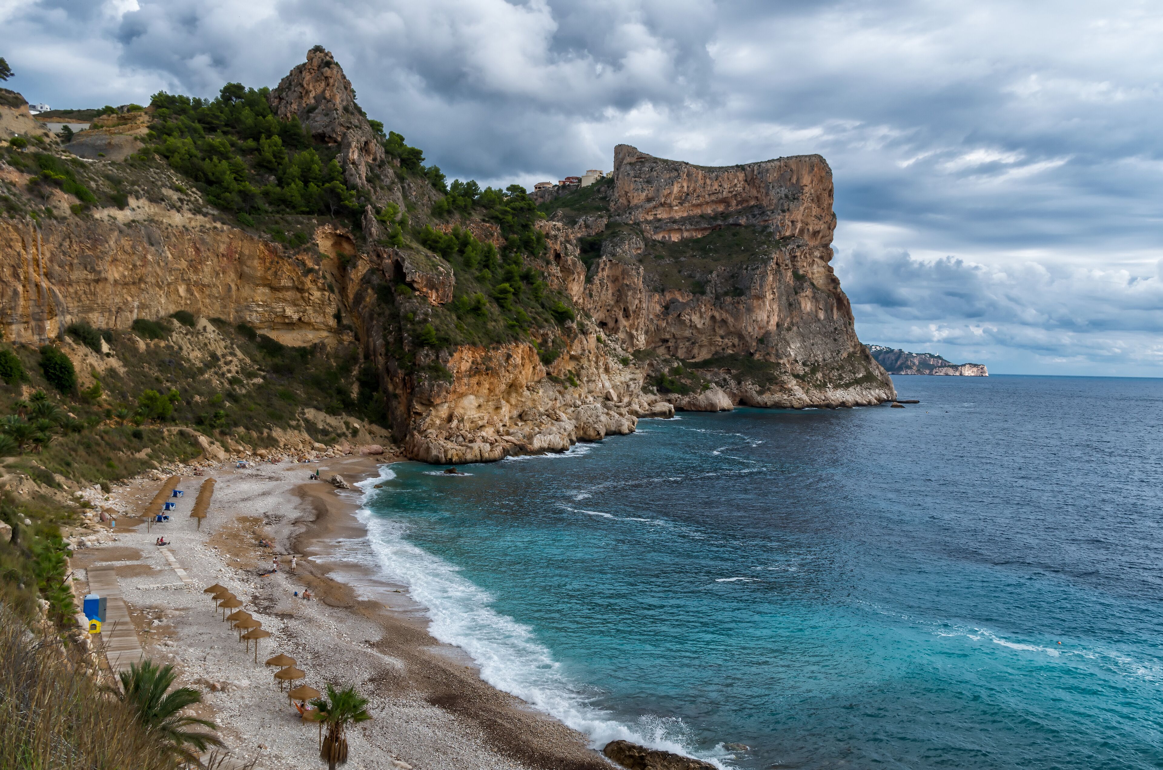 A coastal landscape with a rocky cliff, beach, and clear blue sea.
