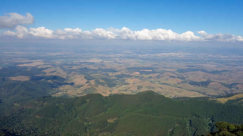 A vast landscape with rolling hills, dense forests, and a clear blue sky with scattered clouds.