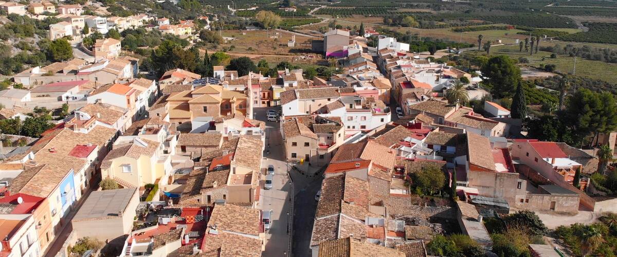 A town with terracotta-roofed buildings, a mountain range in the background, and a clear sky.