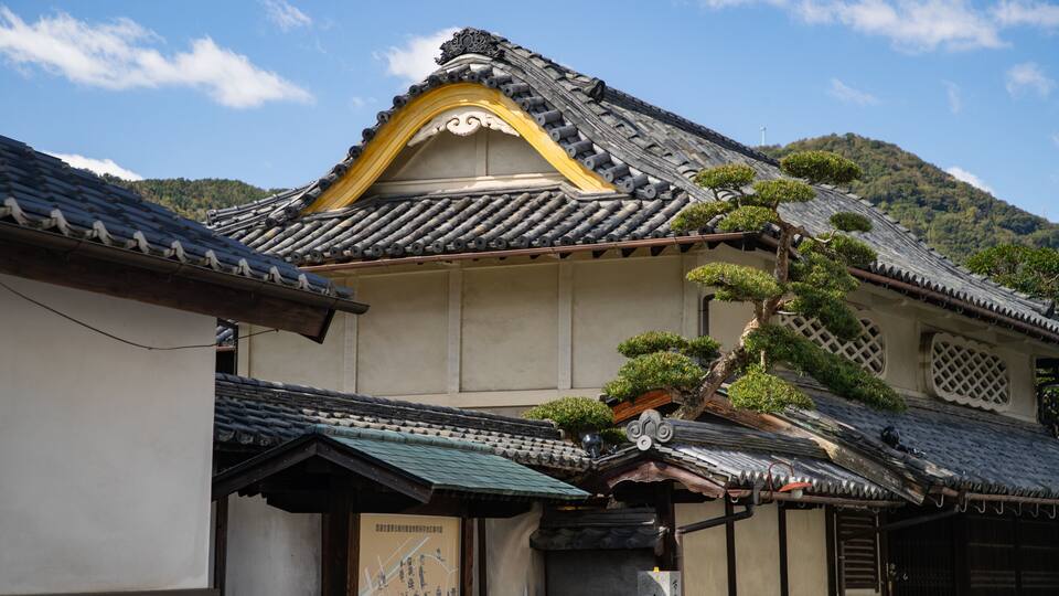 A traditional Japanese building with a tiled roof, a decorative yellow arch, and a bonsai tree.