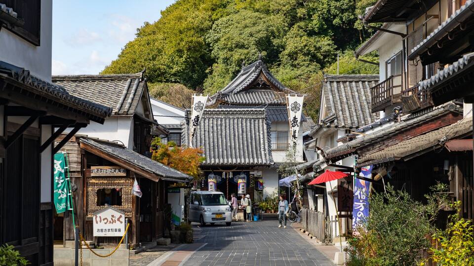 A traditional Japanese street with tiled roofs, wooden buildings, and a car parked in the middle.