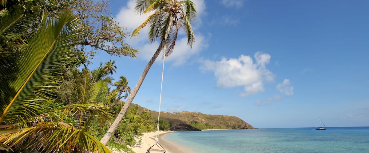A beach with clear turquoise water, a hammock, palm trees, and a boat in the distance.