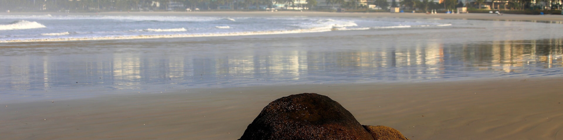 A beach with sand and calm waters, a stone in the foreground and a city in the horizon. Riviera de Sao Lourenco, Bertioga, Brazil