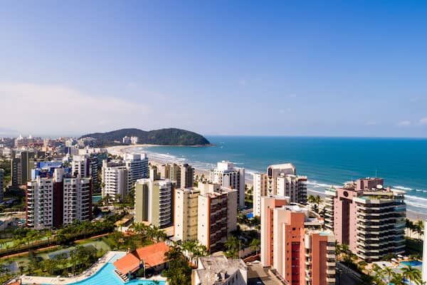 Paisagem costeira com edifícios altos, uma praia e uma montanha ao longe.