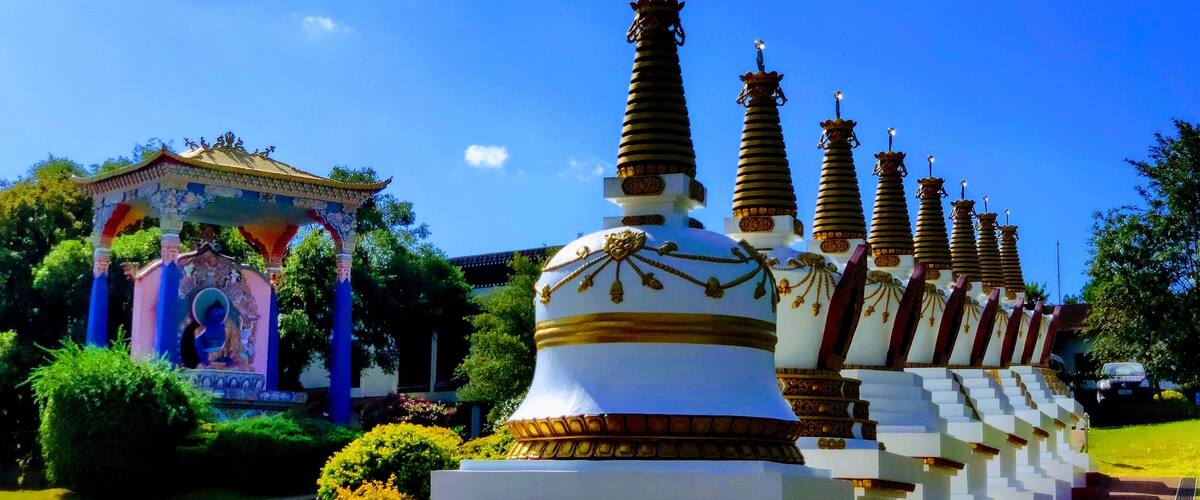 A row of white stupas with golden accents, a colorful pavilion, and lush greenery under a clear blue sky.