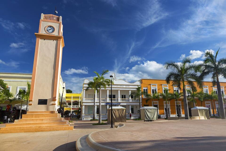 Una torre del reloj, palmeras y edificios históricos bajo un cielo azul.