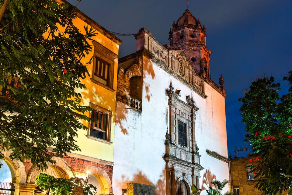 A historic building with a prominent bell tower and arches, surrounded by lush greenery and a well-maintained garden.