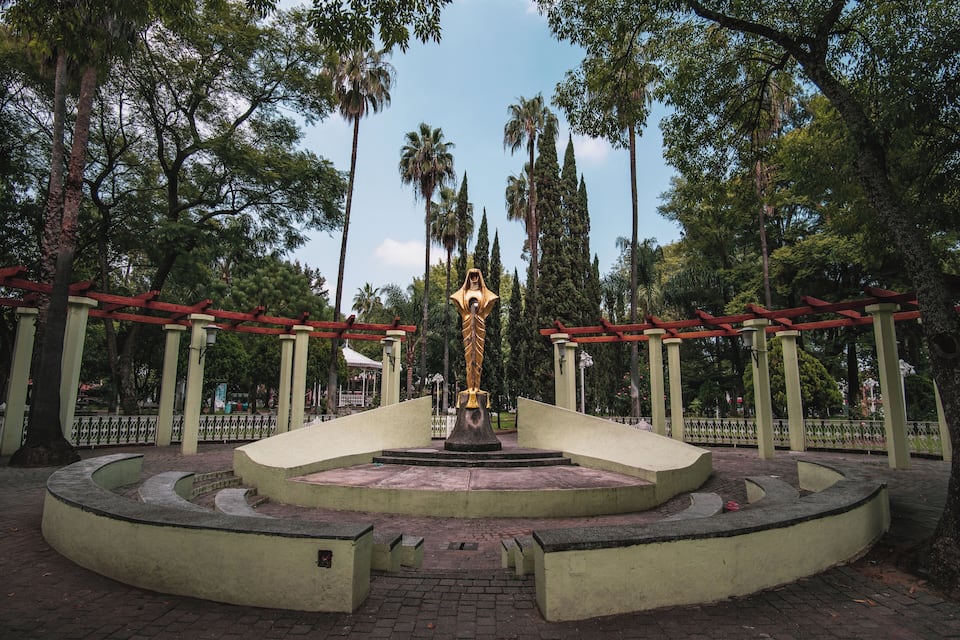 Un parc abritant une statue dorée au centre, entourée d’arbres et de bâtiments aux toits rouges.