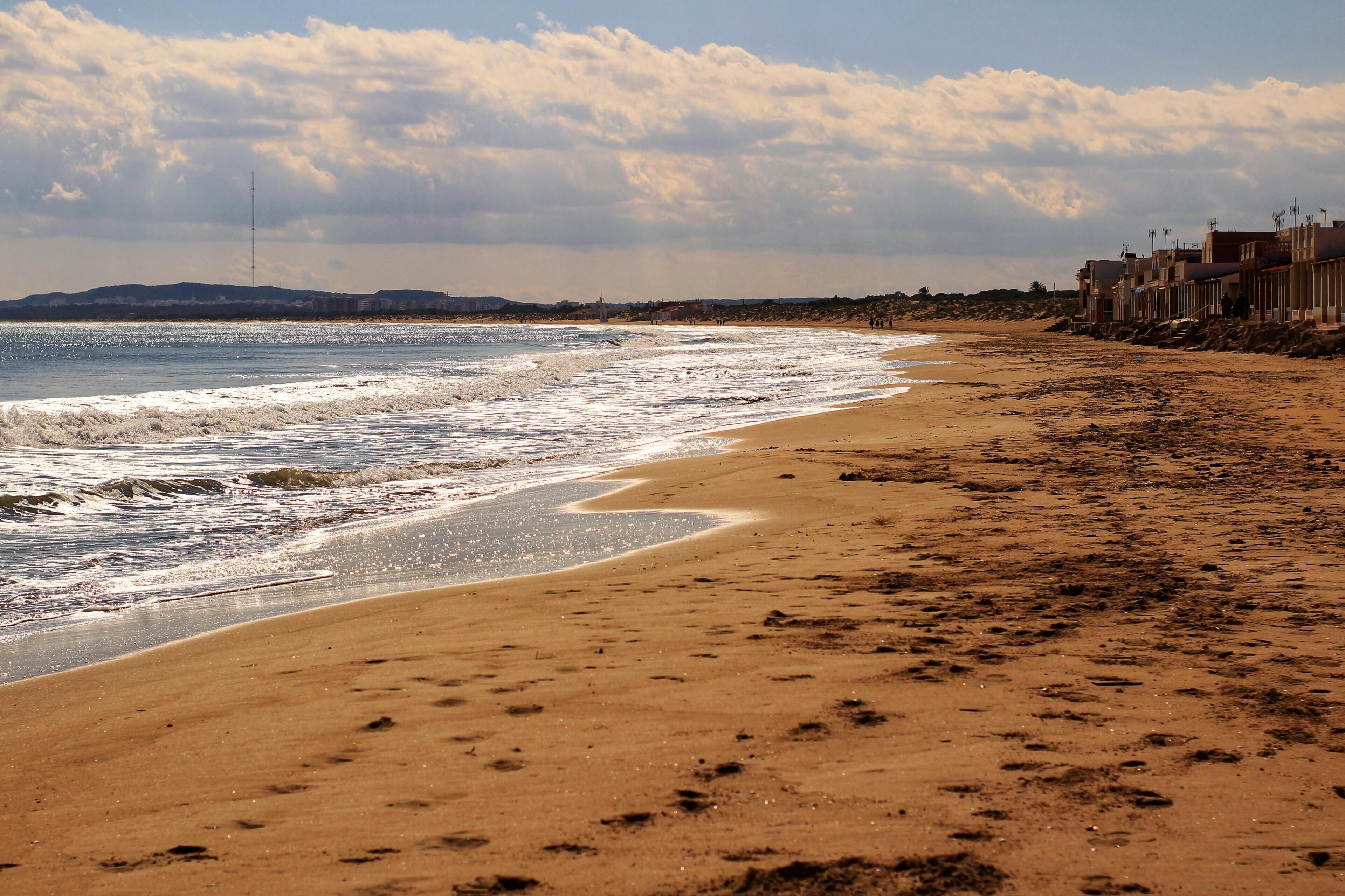 A sandy beach with waves, a cloudy sky, and buildings in the background.