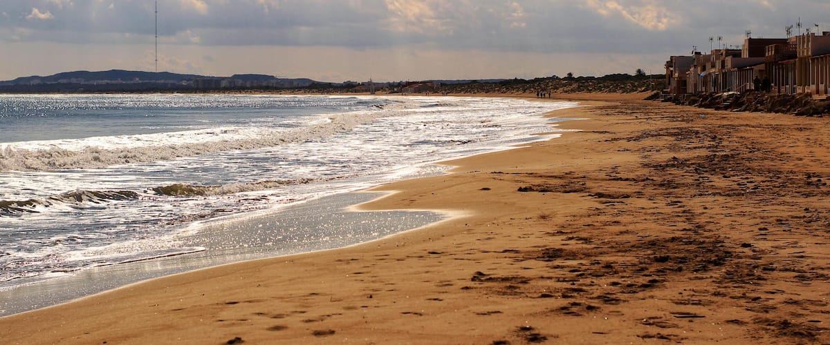 A sandy beach with waves, a cloudy sky, and buildings in the background.