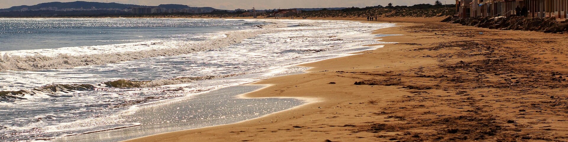 A sandy beach with waves, a cloudy sky, and buildings in the background.