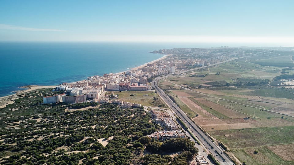 An aerial view of a coastal city with a beach, residential buildings, and agricultural fields.