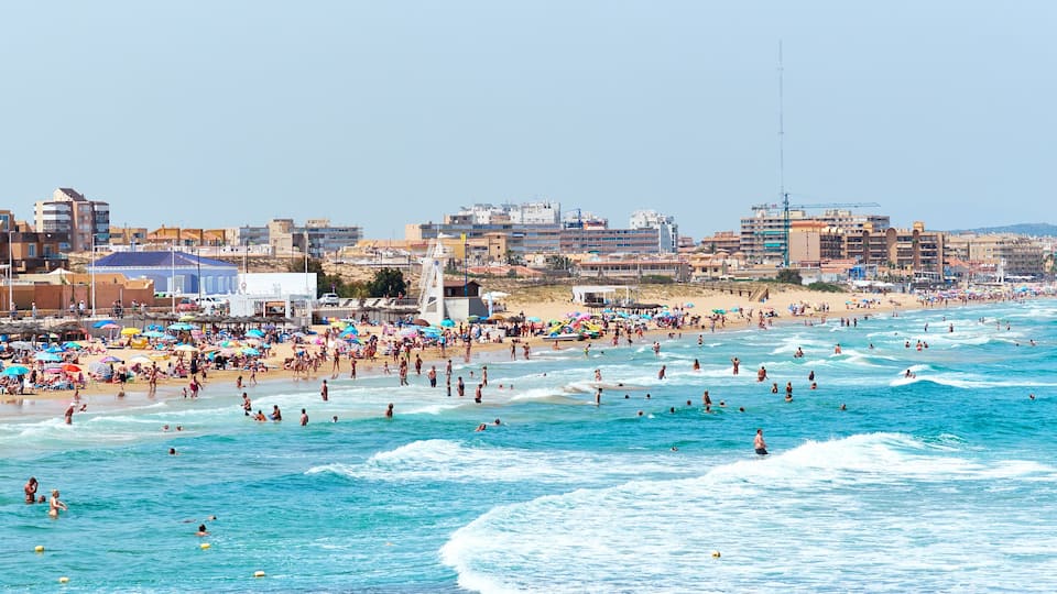 A crowded beach with people swimming and surfing, and buildings in the background.