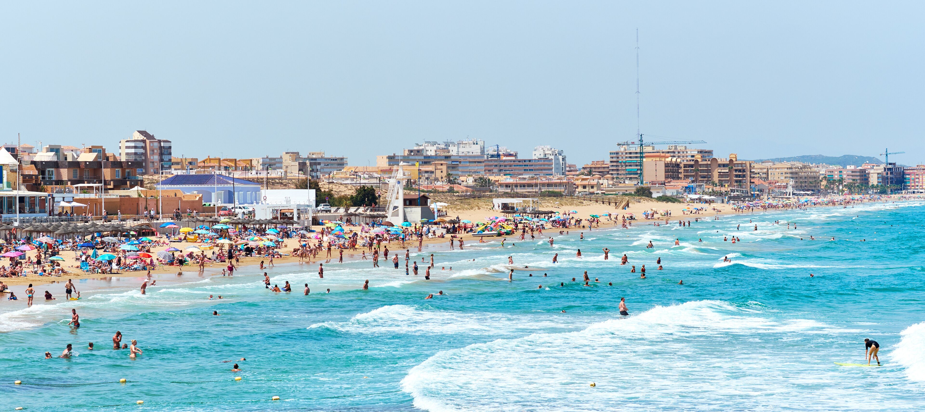 A crowded beach with people swimming and surfing, and buildings in the background.