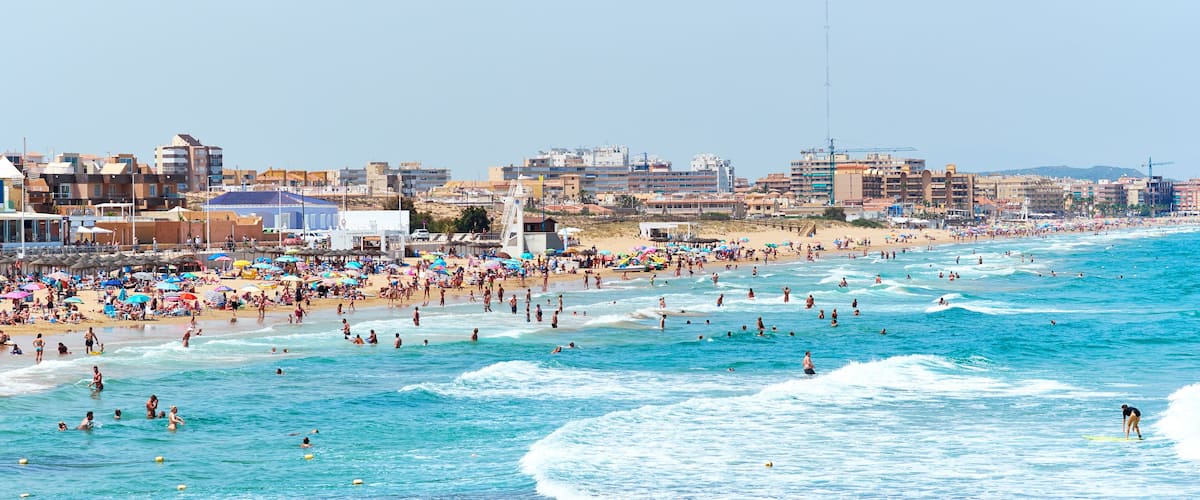 A crowded beach with people swimming and surfing, and buildings in the background.