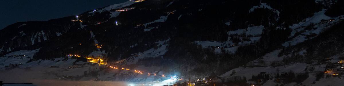 A snow-covered mountain town at night with illuminated buildings and a road leading up the mountain.