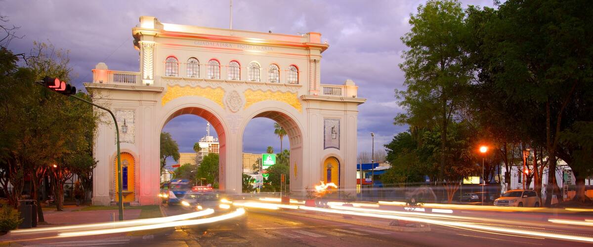 A historic archway with illuminated signs, a busy intersection with light trails from moving vehicles, and surrounding trees.