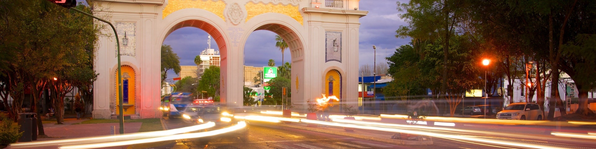 A historic archway with illuminated signs, a busy intersection with light trails from moving vehicles, and surrounding trees.