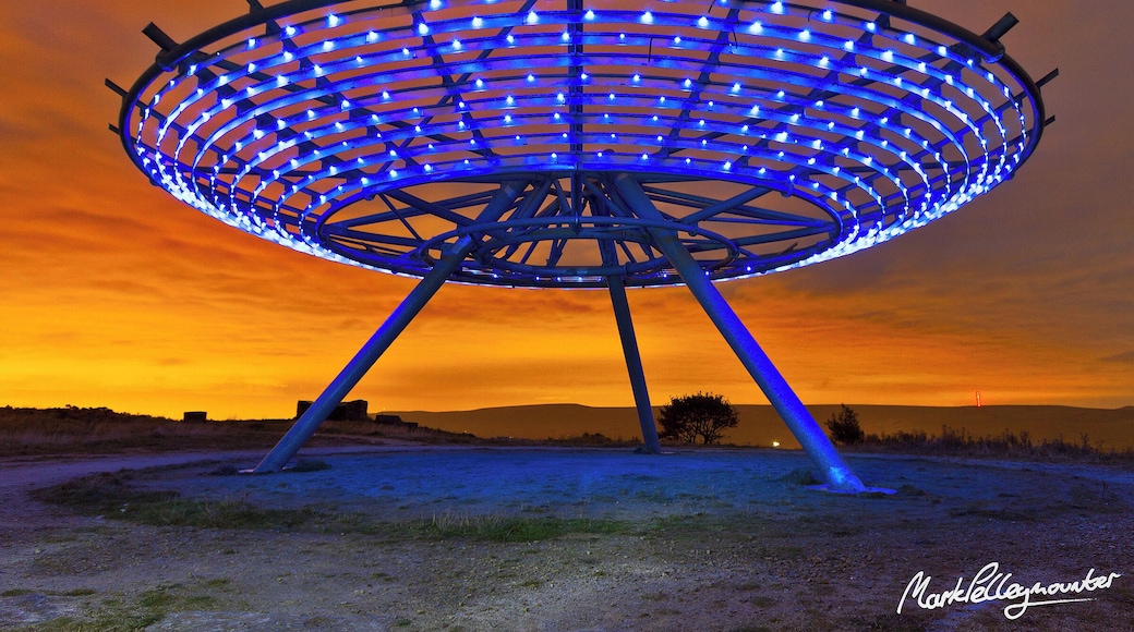 The Halo is an artwork set on the expended landfill site overlooking the town of Haslingden in Rossendale, positioned to be clearly visible from the M66 and A56 approach to Lancashire. It is located at grid reference SD791236. The Halo was the fourth and final Panopticon to be constructed in Lancashire and was launched in September 2007.
The Halo is an 18m-diameter steel lattice structure supported on a tripod five metres above the ground. The core is open at the top, framing views of the sky. It is lit after dark using low-energy LEDs powered by an adjacent wind turbine and glows a sky-blue colour, giving the effect of hovering above the town. It was designed by John Kennedy of LandLab.