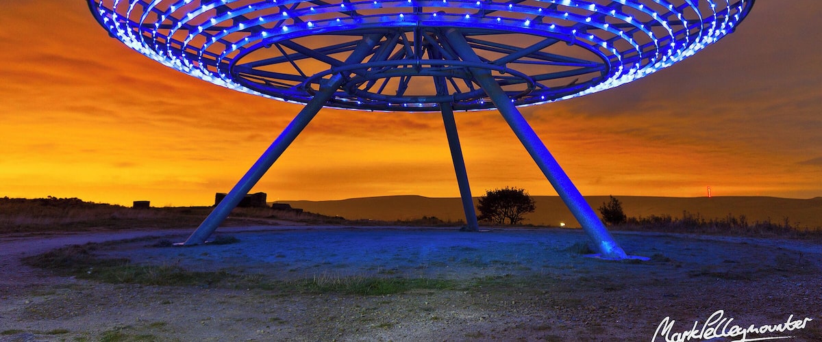 The Halo is an artwork set on the expended landfill site overlooking the town of Haslingden in Rossendale, positioned to be clearly visible from the M66 and A56 approach to Lancashire. It is located at grid reference SD791236. The Halo was the fourth and final Panopticon to be constructed in Lancashire and was launched in September 2007.
The Halo is an 18m-diameter steel lattice structure supported on a tripod five metres above the ground. The core is open at the top, framing views of the sky. It is lit after dark using low-energy LEDs powered by an adjacent wind turbine and glows a sky-blue colour, giving the effect of hovering above the town. It was designed by John Kennedy of LandLab.