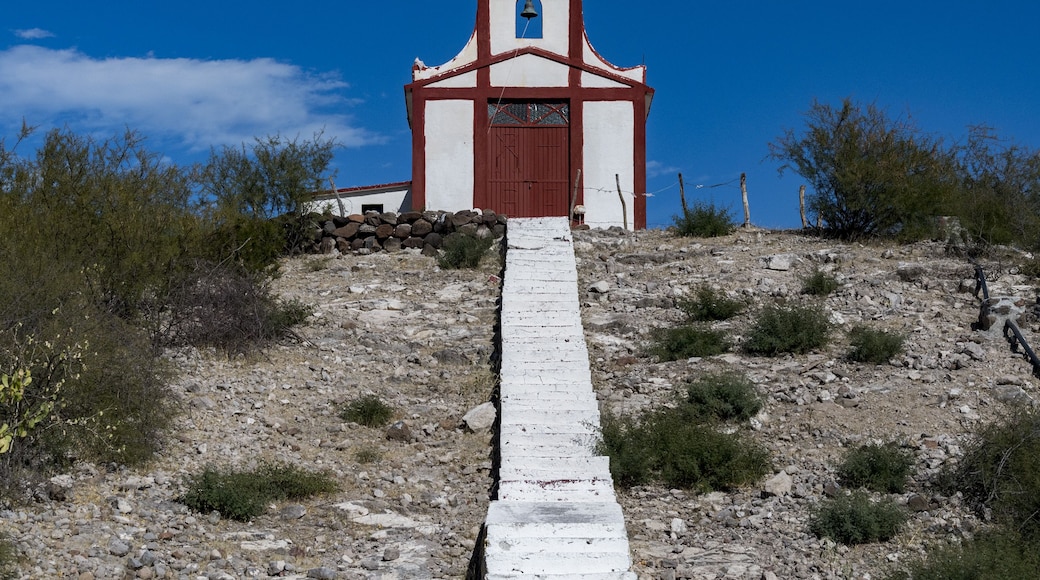 A small church with a white facade and red trim, a bell tower, and a cross on top, situated on a rocky hill with sparse vegetation.