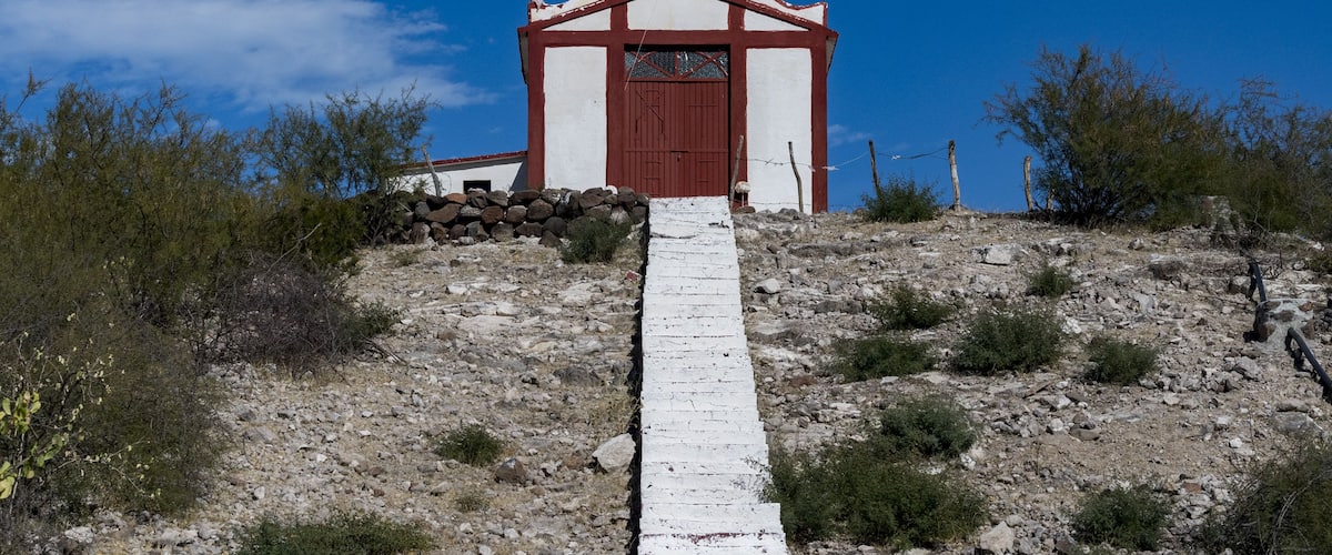 A small church with a white facade and red trim, a bell tower, and a cross on top, situated on a rocky hill with sparse vegetation.