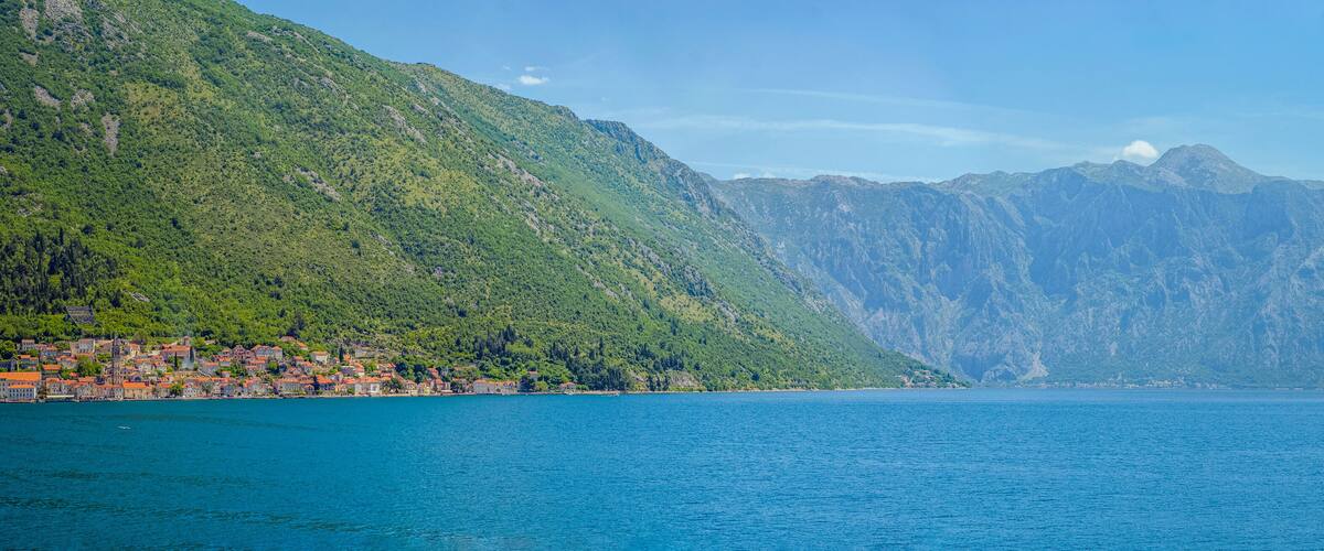 A coastal town with red-roofed buildings, a large body of water, and a mountain range in the background.