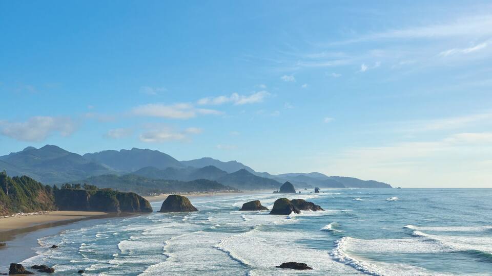 A coastal landscape with a sandy beach, rocky outcrops, and a forested hillside.