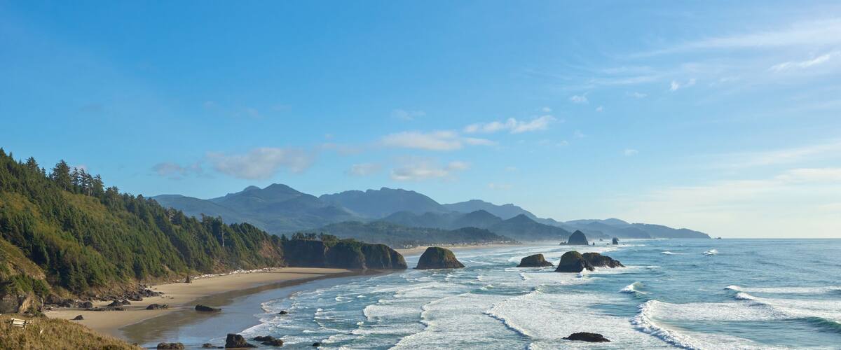 A coastal landscape with a sandy beach, rocky outcrops, and a forested hillside.