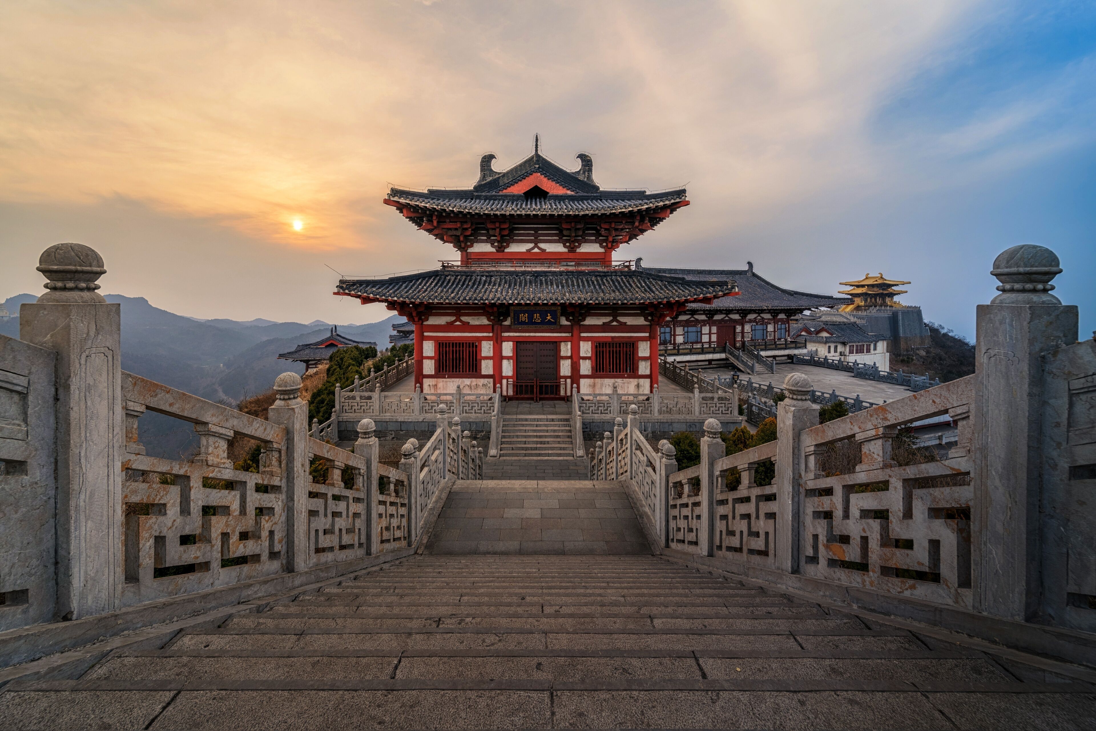 A traditional Chinese building with a multi-tiered roof, a staircase leading up to it, and a mountainous backdrop.