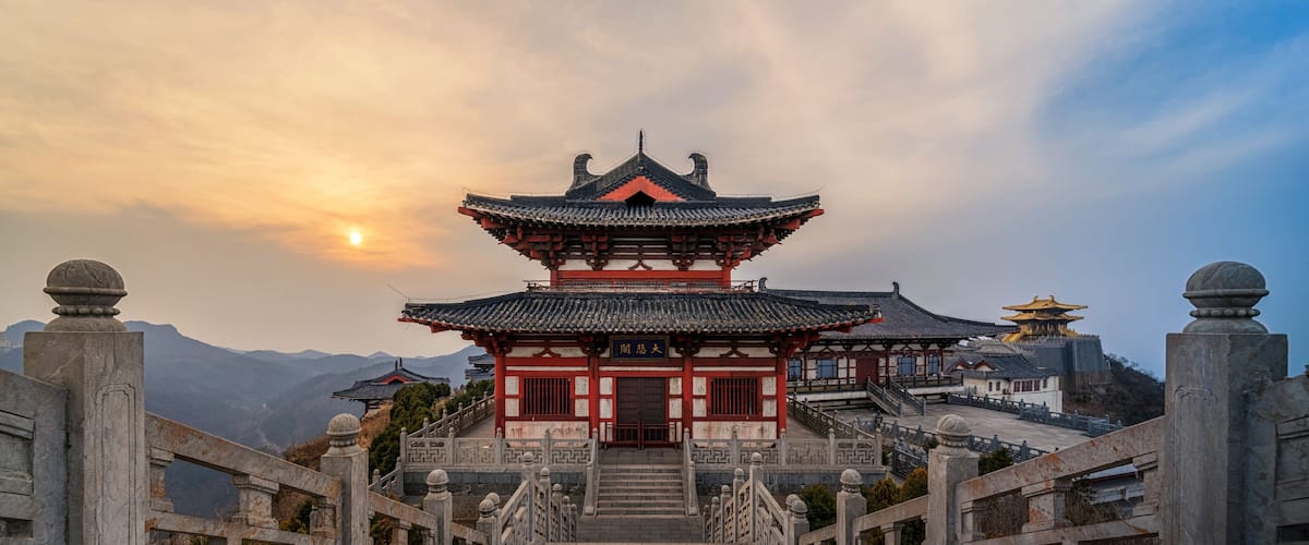 A traditional Chinese building with a multi-tiered roof, a staircase leading up to it, and a mountainous backdrop.