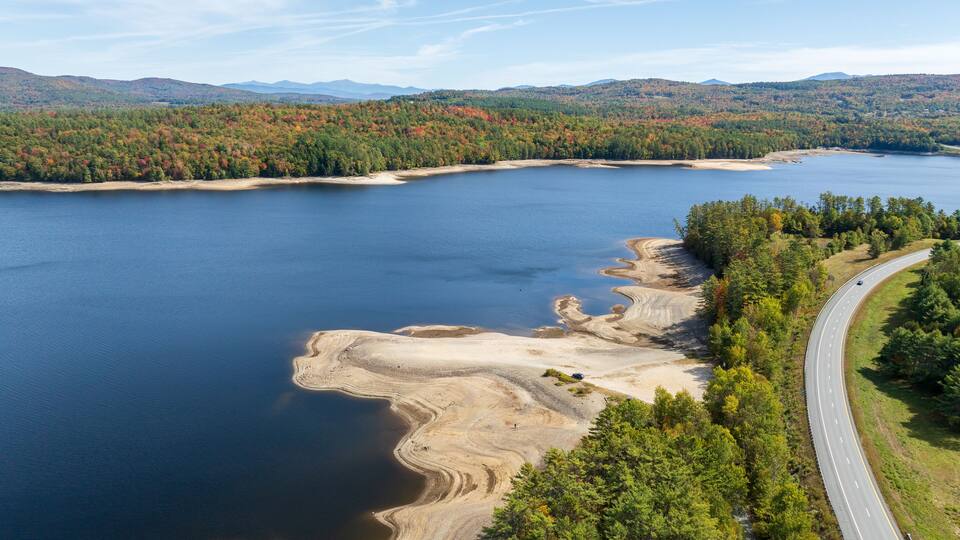 A lake with a winding road alongside, surrounded by autumn-colored forests and distant mountains.