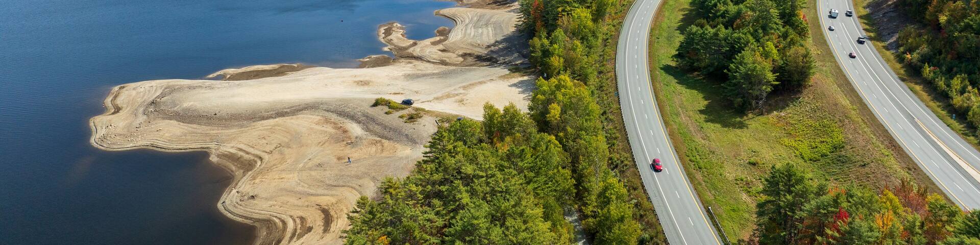 A highway curving alongside a large body of water with forested hills in the background.