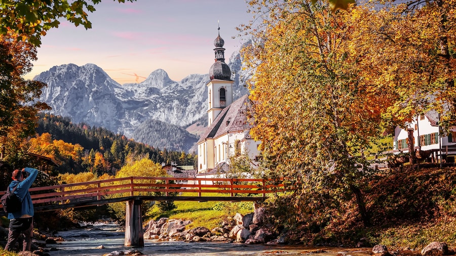Wonderful sunny autumn landscape in Bavarian Alps. Incredible view of famous St. Sebastian Parish Church. Amazing nature scenery with mountains, colorful trees and river. Popular travel destination.