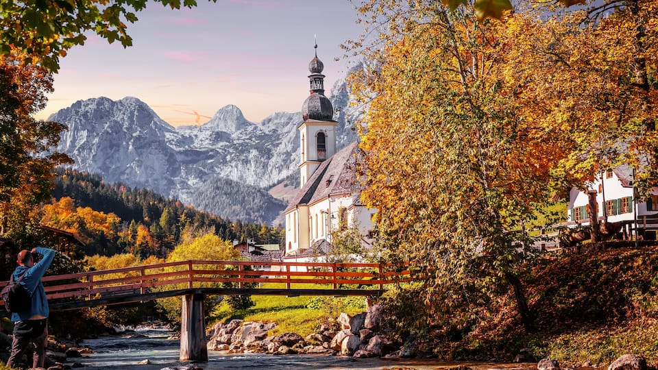 A wooden bridge over a river, a church with a bell tower, and autumn-colored trees in a mountainous valley.