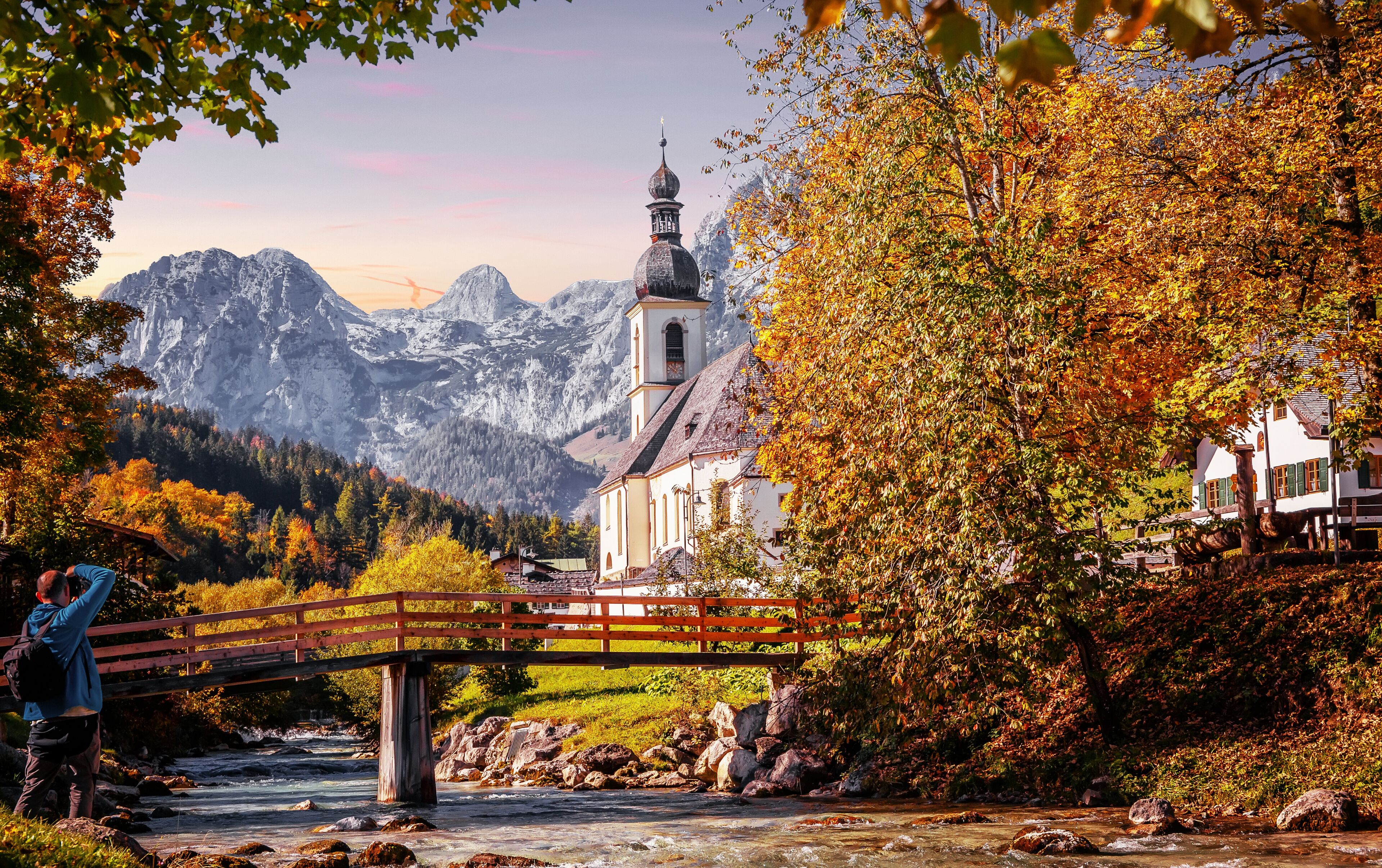 A wooden bridge over a river, a church with a bell tower, and autumn-colored trees in a mountainous valley.