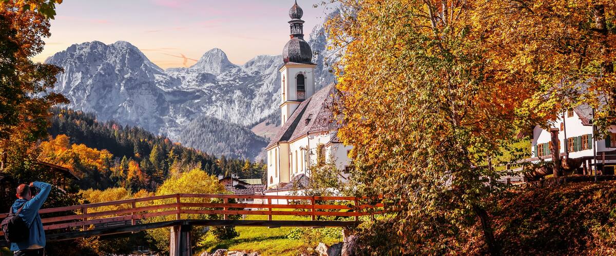 A wooden bridge over a river, a church with a bell tower, and autumn-colored trees in a mountainous valley.