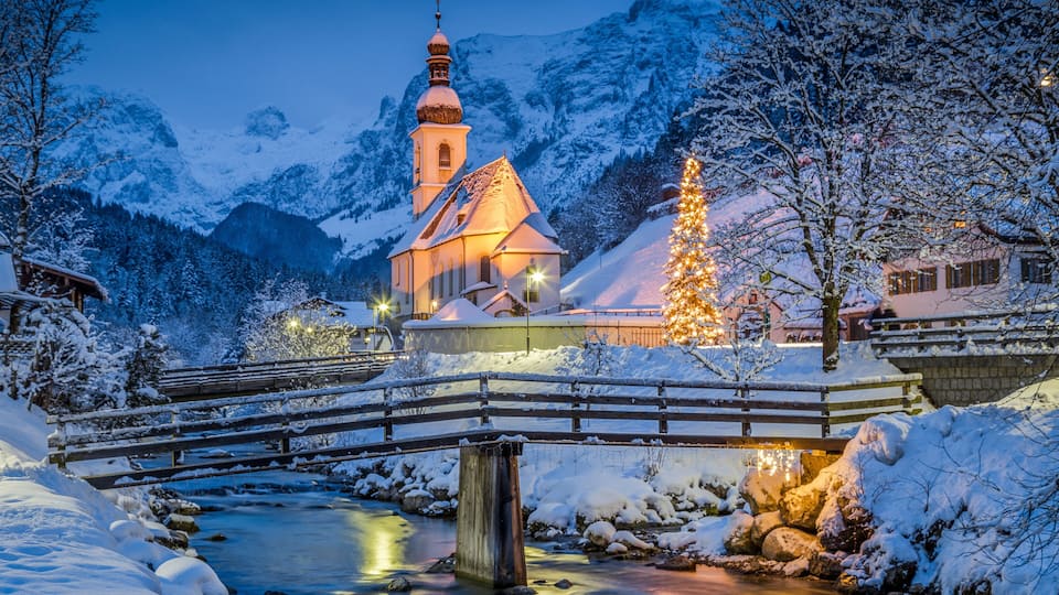 A snow-covered landscape with a church, a bridge, and a Christmas tree.