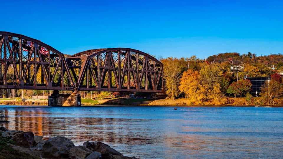 A steel bridge over a calm river, surrounded by autumn foliage and residential buildings.
