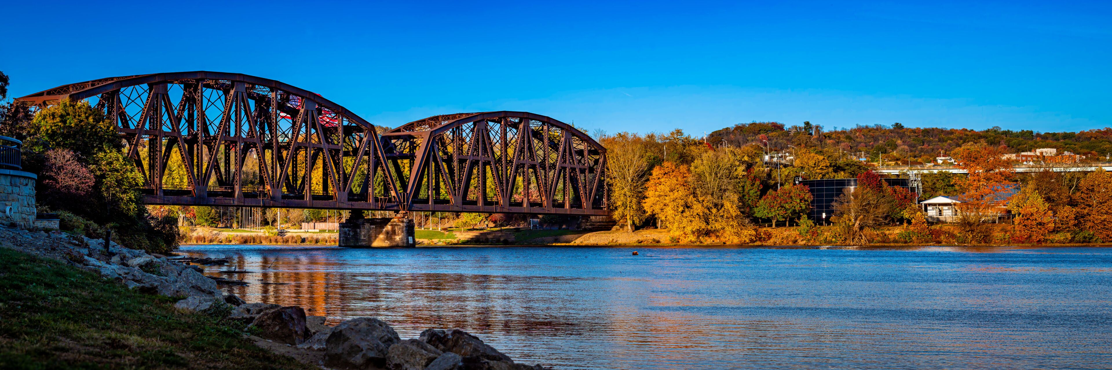A steel bridge over a calm river, surrounded by autumn foliage and residential buildings.