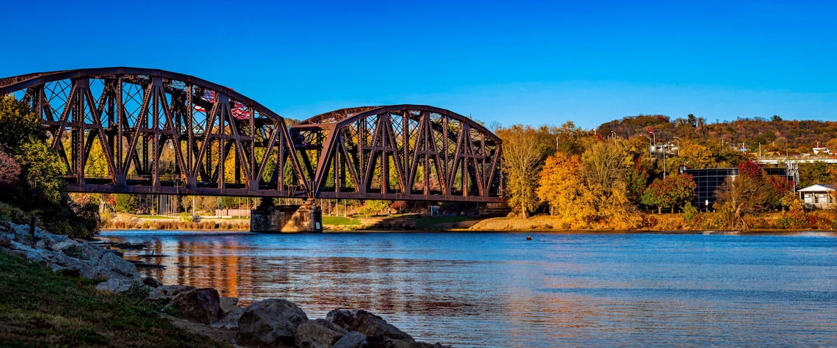 A steel bridge over a calm river, surrounded by autumn foliage and residential buildings.